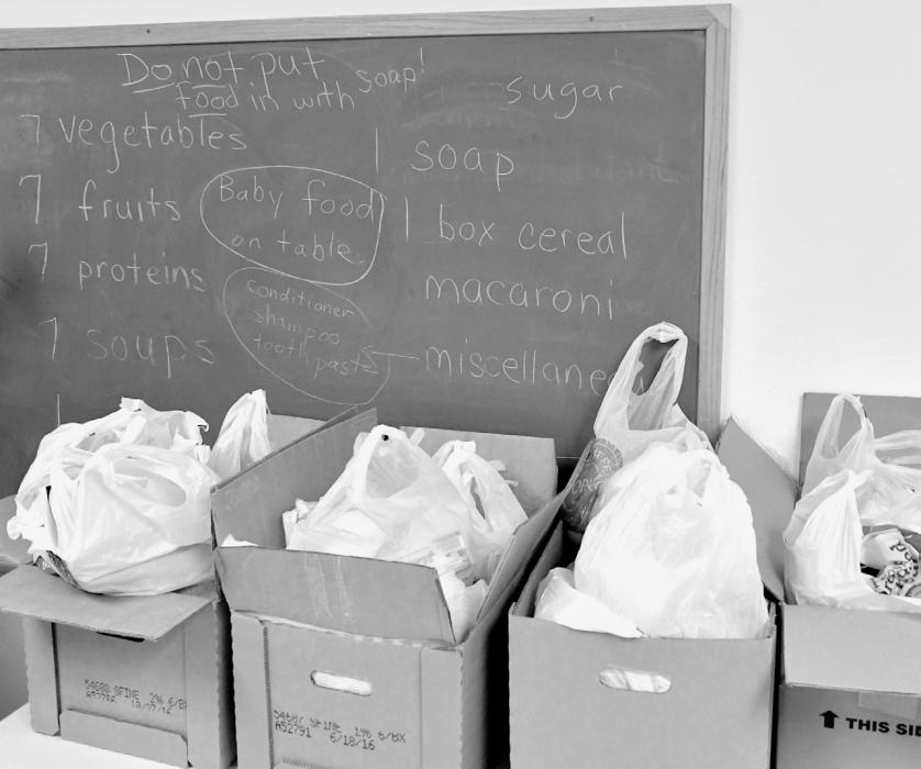 Boxes of food and other items sit waiting for families to pick them up. The chalkboard behind shows what items are put in the boxes, including a balanced combination of vegetables, fruits, proteins, starches and other essentials.